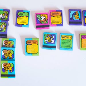 May include: A selection of vintage matchboxes, each with a zodiac sign illustration. The matchboxes have colourful designs and text, including signs like Virgo, Aries, and Sagittarius. The boxes are arranged on a white surface.