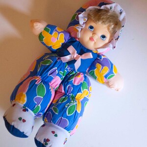 May include: A vintage doll with a soft body and a painted face. The doll is wearing a blue jumpsuit patterned with yellow, pink, and purple flowers. The doll has a matching bonnet and a pink bow at the neck. The doll's feet are white with small red and green floral accents.