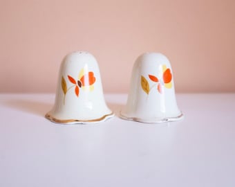 May include: A pair of vintage white ceramic salt and pepper shakers. Each shaker features a bell shape with a scalloped base and a floral design in orange, yellow, and brown. The rims are trimmed with gold and silver.