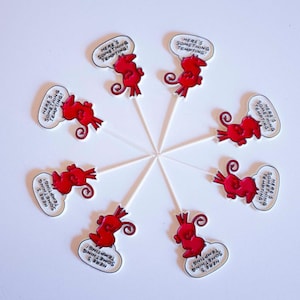 Può includere: Otto decorazioni per cupcake a forma di diavolo rosso disposte in cerchio. Ogni decorazione presenta un diavolo dei cartoni animati con una coda e un fumetto bianco contenente il testo nero "HERE'S SOMETHING TEMPTING!" su un bastoncino bianco.