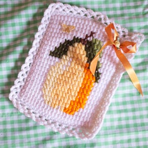 May include: A white, crocheted square with a lemon design. The lemon is yellow and orange with green leaves. A small gold butterfly and an orange ribbon with a bow adorn the square. The background is a green and white checkered pattern.