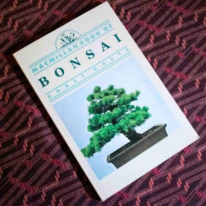 May include: A softcover book titled Macmillan Book of Bonsai by Horst Daute. The white cover features a centered illustration of a green bonsai tree in a rectangular dark gray pot against a light blue background with teal text.