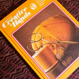 May include: A hardcover book titled Creative Hands with a bright yellow cover. The cover features a graphic of a ball of yellow yarn, a spool of brown thread, a silver thimble, and a pair of silver scissors resting on the yarn.