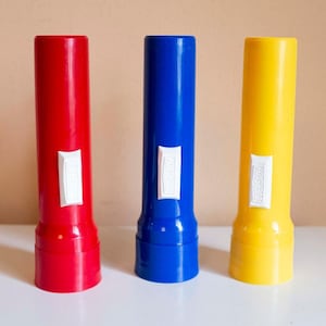 May include: Three colorful flashlights: red, blue, and yellow. Each flashlight has a white rectangular label. The flashlights are cylindrical with a slightly wider base. The background is a neutral beige.