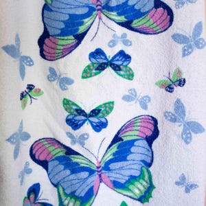 May include: White towel featuring a repeating butterfly pattern. The butterflies are in shades of blue, green, and pink. Smaller, light blue butterfly silhouettes are scattered across the background.