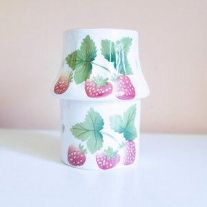 May include: A white ceramic oil burner featuring a two-part design with a flared top section. The exterior is decorated with a repeating pattern of red strawberries and green leaves. Small circular ventilation holes are visible on the side.