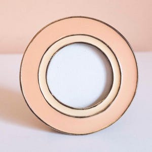 May include: A circular metal pin featuring a wide outer ring of soft peach enamel and a narrower inner ring of cream enamel. The design is framed by thin metallic edges, creating a layered, concentric appearance with a hollow center.