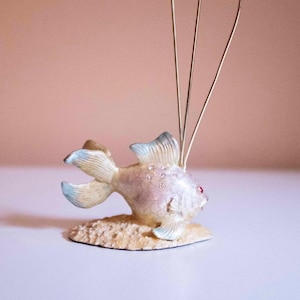 May include: A decorative fish figurine with a light purple body, blue-tipped fins, and a red eye. The fish is embellished with small, clear rhinestones and sits on a textured, beige base. Three thin, gold-colored wires extend upwards from the fish's back.
