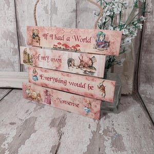 May include: A decorative wooden sign with four stacked planks, featuring the quote "If I had a World of my own, Everything would be Nonsense." The sign has Alice in Wonderland illustrations and a jute rope for hanging.