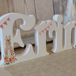 May include: White wooden letters spelling the name "Erin" with a floral and bunny design. The letters have pink and orange floral accents and a string of pink flags.
