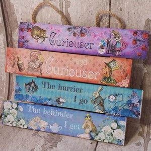 May include: Five wooden signs with whimsical illustrations and text. The signs are stacked on top of each other. The top sign reads "Curiouser and Curiouser". The second sign reads "The hurrier I go". The third sign reads "The behinder I get". The signs are painted in a vintage style with pastel colours and feature characters from Alice in Wonderland.