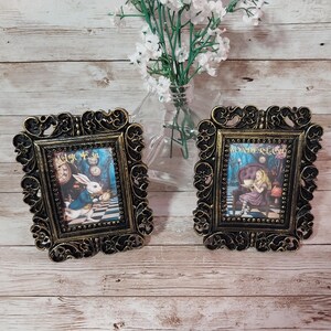 May include: Two decorative picture frames with Alice in Wonderland imagery. The frames are black and gold, with intricate scrollwork. One shows the White Rabbit, the other Alice. The frames are square.