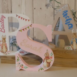 May include: Three wooden letters, painted in pink, white, and beige, spell out the names "Bailey", "Sarah", and "Arlo". Each letter features a small, illustrated rabbit and decorative floral accents.