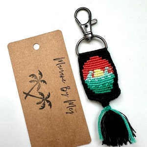 May include: A black and green keychain with a colorful sunset design and a black tassel. The keychain has a silver metal clasp.