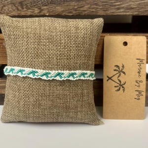 May include: A woven friendship bracelet featuring a repeating teal and white wave pattern. Next to the bracelet is a rectangular brown cardboard tag with a palm tree graphic and the text Macrame By Meg printed in black ink.