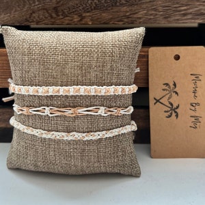 May include: Set of three macrame bracelets in white and beige on a brown burlap pillow. The bracelets are handmade and have a simple, minimalist design. The pillow has a tag that says "Macrame By Meg".
