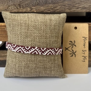 May include: A woven bracelet featuring a repeating geometric pattern in dark red and white thread. The bracelet is wrapped around a square, textured tan burlap pillow. A rectangular kraft paper tag with a palm tree logo and the text Macrame By Meg.