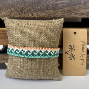 May include: A woven friendship bracelet featuring a repeating wave pattern in shades of teal, light green, and white, with a thin orange border. Beside it is a rectangular brown paper tag with two palm tree icons and the text Marine By Meg.