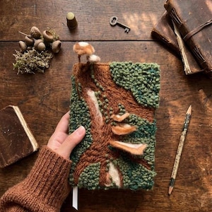 Op de afbeelding: Een handgemaakt notitieboek met een bosscène-ontwerp. De omslag heeft een getextureerde, bruine boomstam met groene bladeren en bruine paddenstoelen. Een sleutel, vingerhoed, eikels en een potlood liggen op het houten oppervlak. Een hand houdt het notitieboek vast.