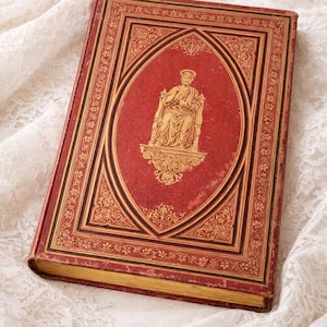 May include: A vintage, hardback book with a red cover and gold embossed detailing. The cover features an oval design with a seated figure and ornate borders. The book's edges are gold, and it rests on a white lace surface.