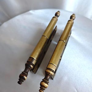 Könnte beinhalten: Zwei antike Messing-Scharniere mit dekorativem Design. Die Scharniere haben einen zylindrischen Körper und eine dekorative Verzierung am Ende. Das Messing hat eine warme, gealterte Patina. Diese Scharniere sind wahrscheinlich für einen Schrank oder eine Tür.