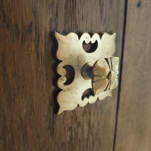 Può includere: Primo piano di una maniglia in ottone con un design floreale. La maniglia è montata su una porta di legno scuro. La maniglia ha una piastra posteriore quadrata con ritagli decorativi.