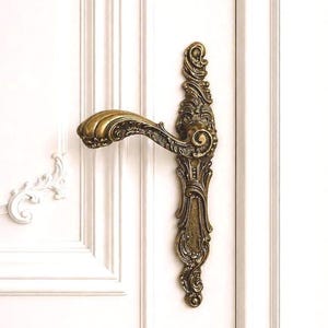 May include: An antique brass door handle featuring detailed scrollwork and a curved lever. The handle is mounted on a white panelled door, displaying a classic, vintage design. The handle's finish has a warm, aged appearance.