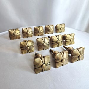 Może przedstawiać: Kolekcja kwadratowych, mosiężnych gałek do szafek w stylu antycznym z kwiatowym wzorem. Gałki ułożone są na białej powierzchni, prezentując swoje postarzone, złote wykończenie. Idealne do renowacji mebli.