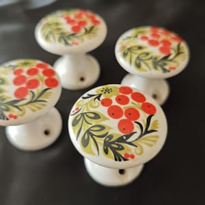 Peut inclure: Quatre boutons en céramique blanche avec un motif floral composé de baies rouges et de feuilles vertes. Les boutons ont une forme simple et ronde et un petit trou au centre pour le montage.