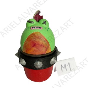 May include: A hand-painted, whimsical frog figurine. The frog is green with a red and orange belly, a black collar with silver spikes, and a red base. The frog has a mohawk and a cross-eyed expression. A tag reads "M1".