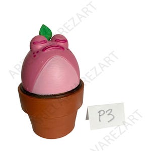 May include: A pink frog-shaped sculpture with closed eyes and a green leaf on top, set in a brown flower pot. The frog has a light pink belly and darker pink details. A small white tag with the text "P3" is visible.