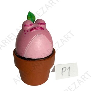 May include: A whimsical, hand-painted pink frog figurine with closed eyes and a green leaf, sitting in a brown flower pot. The frog has a light pink belly and darker pink accents. A small white tag with "P1" is visible.