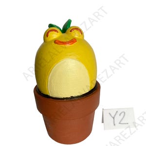 May include: A whimsical, yellow frog-like figurine with a smiling face and green leaves, set in a brown terracotta pot. The figurine has a light yellow belly. A small white tag with the text "Y2" is visible.