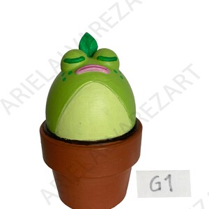 May include: A whimsical, hand-painted ceramic frog figurine in a terracotta pot. The frog is a vibrant green with a white belly, closed eyes, and a small green leaf on its head. The pot is a classic brown, and a small white tag with "G1" is visible.
