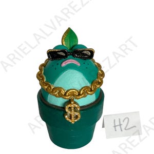 May include: A whimsical, hand-painted ceramic frog figurine in a green pot. The frog wears black sunglasses, a gold chain with a dollar sign pendant, and a gold leaf on its head. The pot is dark green, and a small white tag with "H2" is visible.