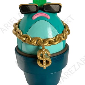 May include: A whimsical teal-colored cactus figurine wearing black sunglasses and a gold chain with a dollar sign pendant. The cactus is in a dark teal pot, with a green leaf on top.