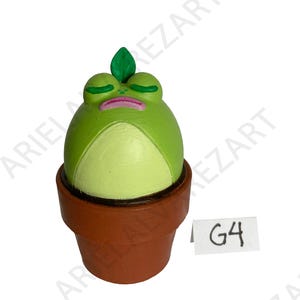 May include: A whimsical, hand-painted frog figurine in a terracotta pot. The frog is light green with a lighter green belly, closed eyes, and a pink mouth. A green leaf sprouts from its head. A small white tag reads "G4".