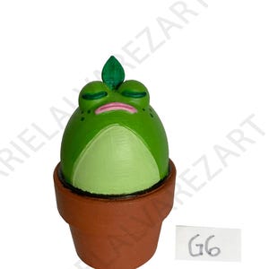 May include: A whimsical, hand-painted frog figurine with closed eyes and a leaf on its head, sitting in a terracotta pot. The frog is primarily green with lighter green accents and a pink mouth. The pot has a brown color and the tag says G6.