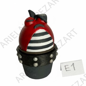 May include: A whimsical, red, egg-shaped figurine with a black and white striped torso, a black hat, and a studded black base. The figurine has a cartoonish face and is labeled "E1" on a small white tag.