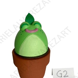 May include: A whimsical, green frog-shaped figurine with a leaf on its head, set in a brown terracotta pot. The frog has a smiling face with pink lips and white belly. A small white tag with the text "G2" is visible.