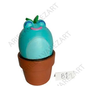 May include: A whimsical, turquoise frog figurine with a smiling face, blue eyes, and green leaves, sits in a brown terracotta pot. The frog has a white belly and pink mouth. A small white tag with the text "B1" is visible.