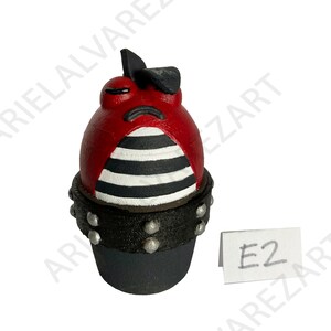 May include: A red and black decorative figurine with a sad face, black bow, and black and white striped midsection. The base is black with silver studs. A small white tag with "E2" is visible.