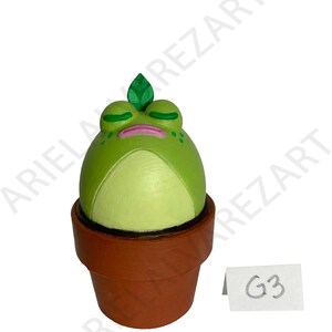 May include: A whimsical, hand-painted frog figurine in a terracotta pot. The frog is a vibrant green with closed eyes, a pink mouth, and a small green leaf on its head. The pot is a classic brown color. A small white tag with "G3" is visible.