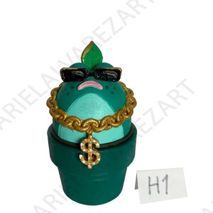 May include: A whimsical, hand-painted figurine of a plant wearing sunglasses and a gold chain with a dollar sign pendant. The plant is in a green pot, with a green leaf on top. The figurine has a sad expression.