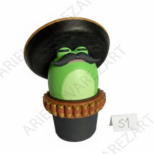 May include: A whimsical green frog figurine wearing a black sombrero with a gold trim and a large black mustache. The frog has a pink mouth and is perched atop a black base with a brown and gold decorative band.