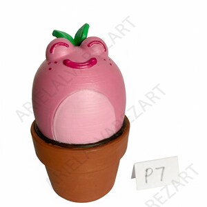 May include: A pink frog-shaped figurine with a smiling face and green leaves on top, sitting in a brown terracotta pot. The figurine has a light pink belly and small dark pink spots. A small white tag with the text "P7" is next to the pot.