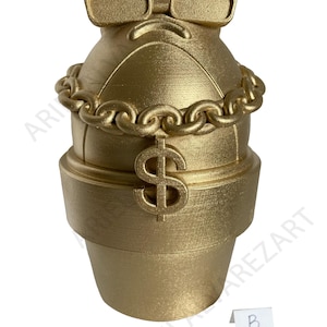 May include: A gold-colored decorative sculpture resembling a character with sunglasses and a chain necklace. A dollar sign is featured on the front. The sculpture is placed on a white background.