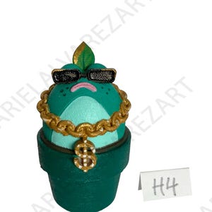 May include: A whimsical, green frog figurine wearing black sunglasses and a gold chain with a dollar sign pendant. The frog is in a green pot, with a leaf on its head. A small white tag with "H4" is visible.