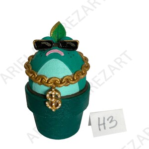 May include: A whimsical, hand-painted ceramic cactus figurine in a green pot. The cactus has a turquoise face, black sunglasses, and a gold chain with a dollar sign pendant. A small white tag with "H3" is visible.