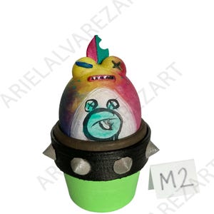 May include: A whimsical, hand-painted ceramic frog sculpture. The frog features a rainbow-colored head, a black studded collar, and a green base. The frog's face has a cartoonish design with a turquoise mouth and blue eyes. The sculpture has the text "M2" on a small white tag.